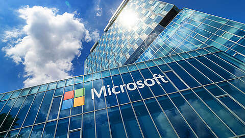 Ein modernes gläsernes Bürogebäude mit dem Microsoft-Logo und einem bunten Symbol auf der Fassade, in dem sich der blaue Himmel und die Wolken darüber spiegeln.