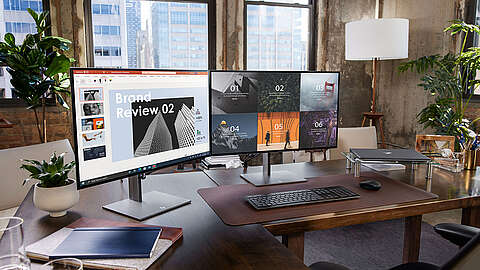 Ein moderner Bürotisch mit zwei großen Monitoren, auf denen Präsentationsfolien zu sehen sind, einer Tastatur, einer Maus, einem Laptop, einer Schreibtischlampe, Topfpflanzen und großen Fenstern, aus denen die Gebäude der Stadt zu sehen sind.