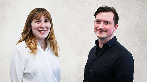 Zwei Personen stehen nebeneinander vor einem hellgrauen Hintergrund, die eine trägt ein weißes Hemd, die andere ein schwarzes Hemd, beide lächeln in die Kamera.