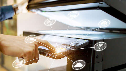 Eine Person drückt eine Taste an einem digitalen Drucker oder Kopierer. Weiße, leuchtende Netzwerksymbole, die Konnektivitätsfunktionen wie Wi-Fi, Cloud und Drucken darstellen, sind um das Bedienfeld herum eingeblendet.