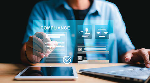 Eine Person interagiert mit digitalen Compliance-Symbolen auf einem transparenten Bildschirm, mit einem Tablet und einem Laptop auf einem Schreibtisch, die gesetzliche Vorschriften und geschäftliche Compliance-Prozesse symbolisieren.