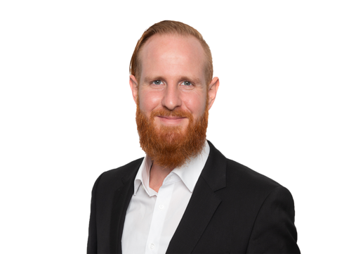 Ein Mann mit roten Haaren und Bart, der ein schwarzes Jackett und ein weißes Hemd trägt, lächelt vor einem weißen Hintergrund in die Kamera.