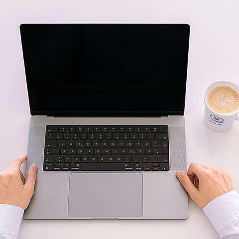 Ein leerer Laptop-Bildschirm auf einem Holztisch neben einer Tasse Kaffee, einem Grafiktablett, einem Notizblock und einer Topfpflanze. Die Umgebung vermittelt den Eindruck einer gemütlichen Arbeitsumgebung.