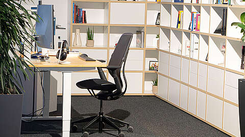 Ein modernes Büro mit einem Schreibtisch, einem Computer, Kopfhörern und einem Bürostuhl. Regale mit Büchern und Pflanzen säumen den Hintergrund. Der Raum ist aufgeräumt und gut beleuchtet, mit einer großen Pflanze auf der linken Seite.