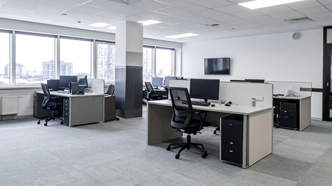 Ein modernes Großraumbüro mit mehreren leeren Schreibtischen, Bürostühlen, Computern und großen Fenstern, die natürliches Licht hereinlassen. Der Raum ist sauber und hell, die Möbel sind in neutralen Farben gehalten und der Boden ist mit Teppich ausgelegt.