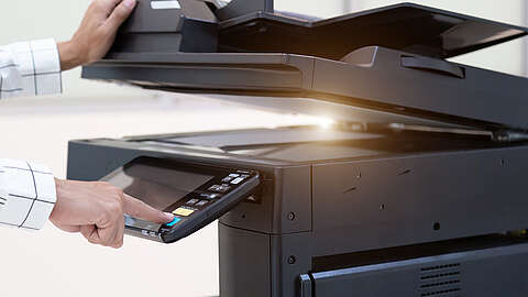 Eine Person bedient das Bedienfeld eines Schwarzkopierers oder Multifunktionsdruckers, indem sie mit einer Hand eine Taste drückt und mit der anderen die obere Klappe öffnet.