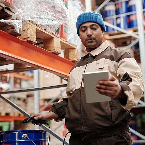 Ein Arbeiter in braun-beiger Uniform und blauer Mütze scannt in einem Lager mit einem Tablet Artikel mit einem Barcode-Scanner. Im Hintergrund sind Regale mit großen Behältern und Kisten zu sehen.