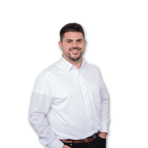 Ein Mann mit kurzen dunklen Haaren und Bart, der ein weißes Button-up-Hemd und eine dunkle Hose trägt, steht vor einem schlichten weißen Hintergrund und lächelt, eine Hand in der Tasche.